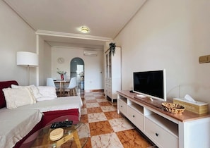 A living room with a sofa, TV, coffee table, and a dining area in the background.