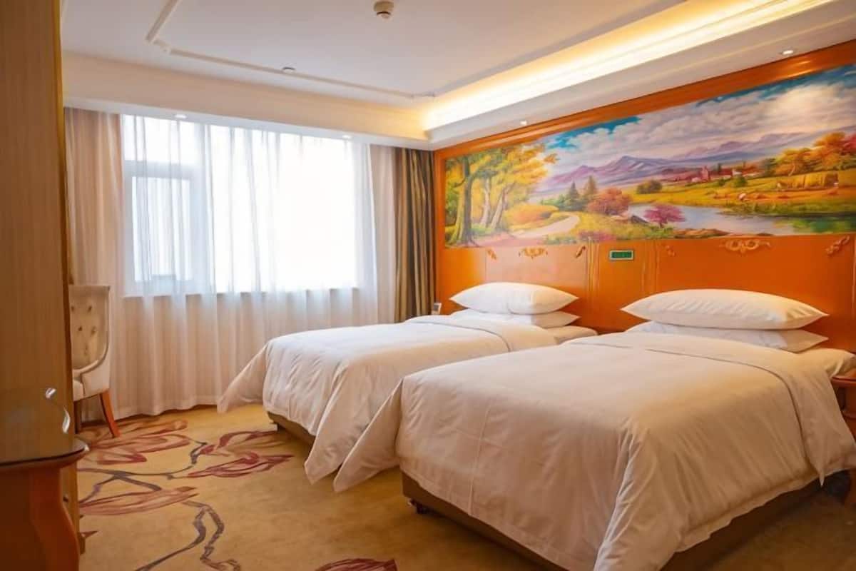A hotel room with two beds, a painting on the wall, and a window with curtains.