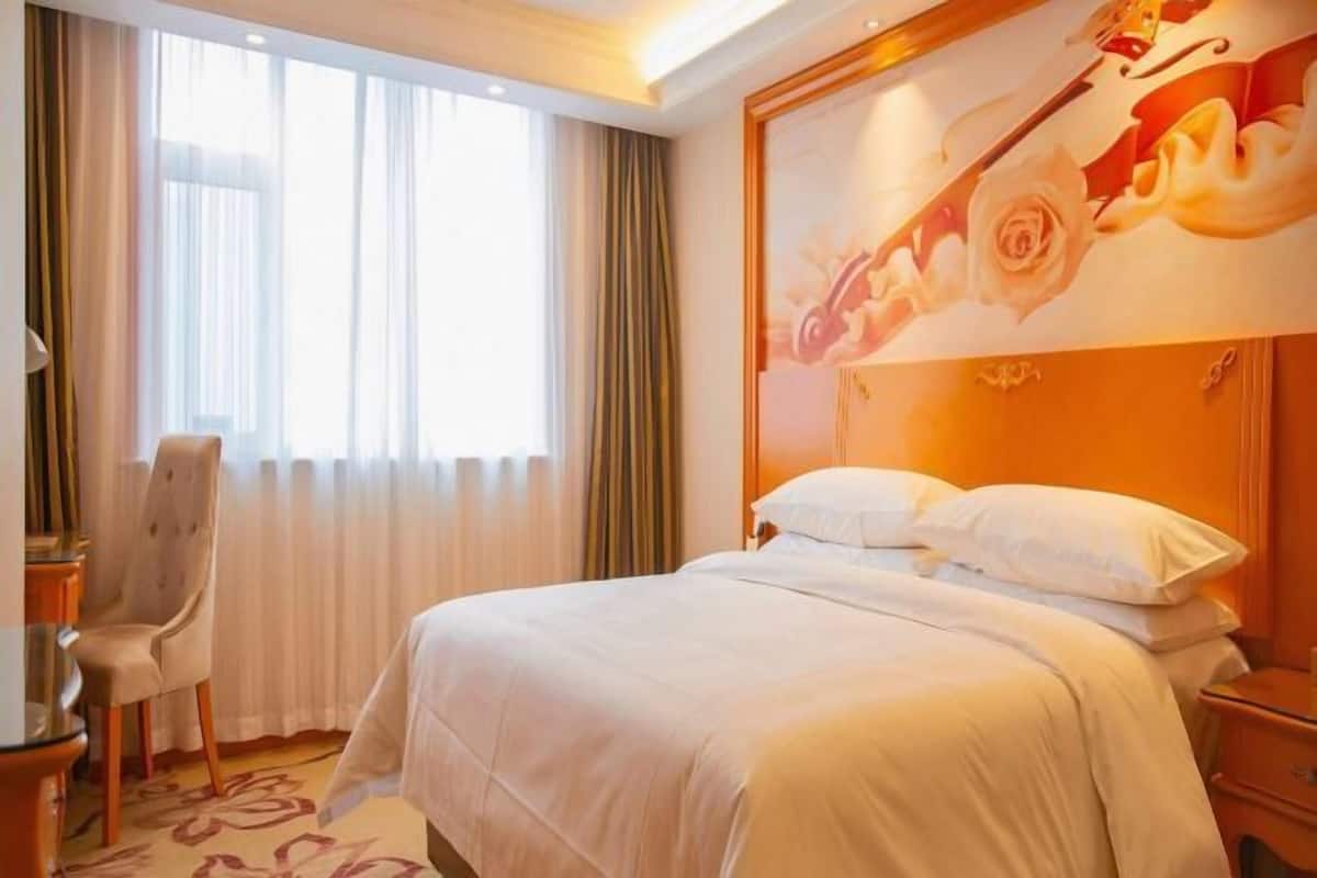 A hotel room with a bed, a chair, a nightstand, and a large window with curtains.