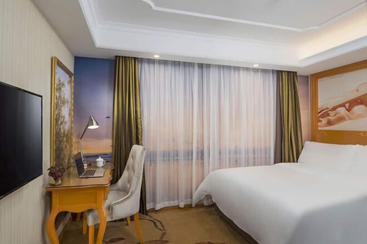 A hotel room with a bed, a desk with a laptop, a chair, a television, and a large window with curtains.