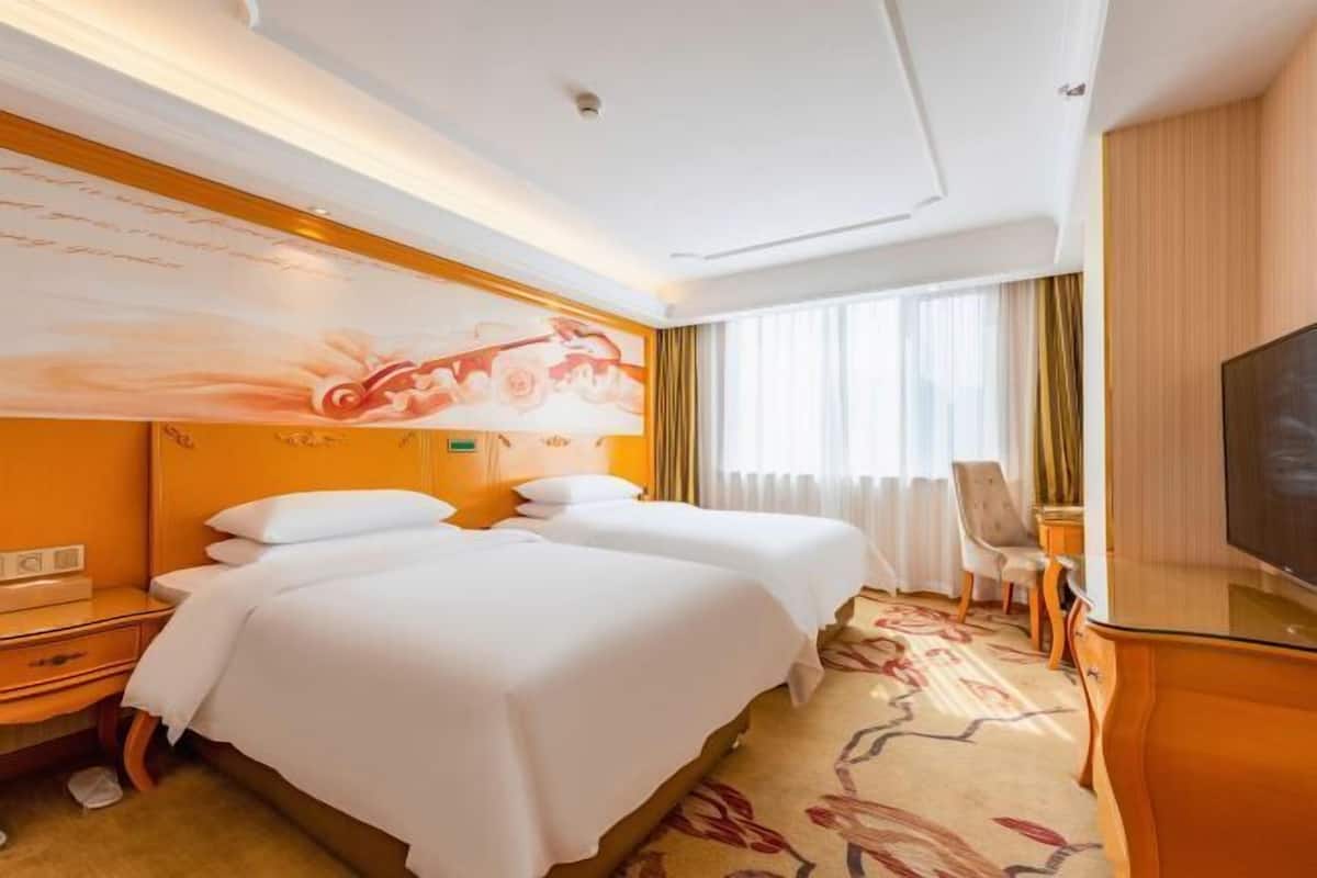 A hotel room with two beds, a desk, a chair, a television, and a large window with curtains.