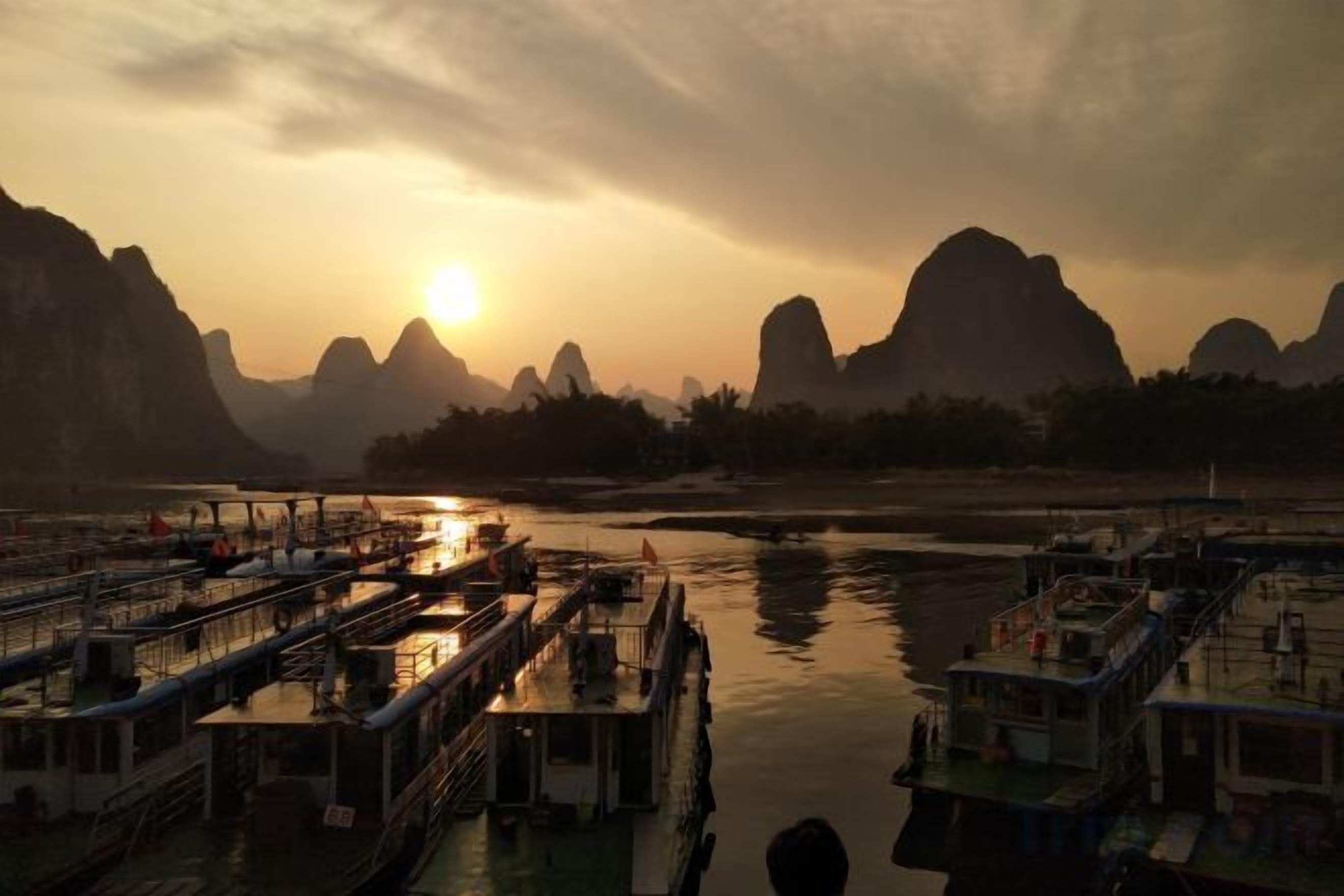 A riverfront with boats docked, a bridge, and mountains in the background.