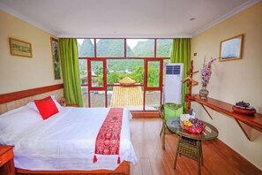 Habitación de hotel con cama, una mesita con fruta, una silla, una ventana con vista a un templo y un estante con adornos.