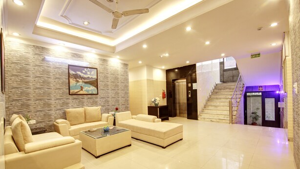 A spacious lobby with a modern design, featuring a large seating area with sofas and a coffee table, a staircase leading to an elevator, and a wall-mounted painting. - Hotel Vibe Blue (Noida)