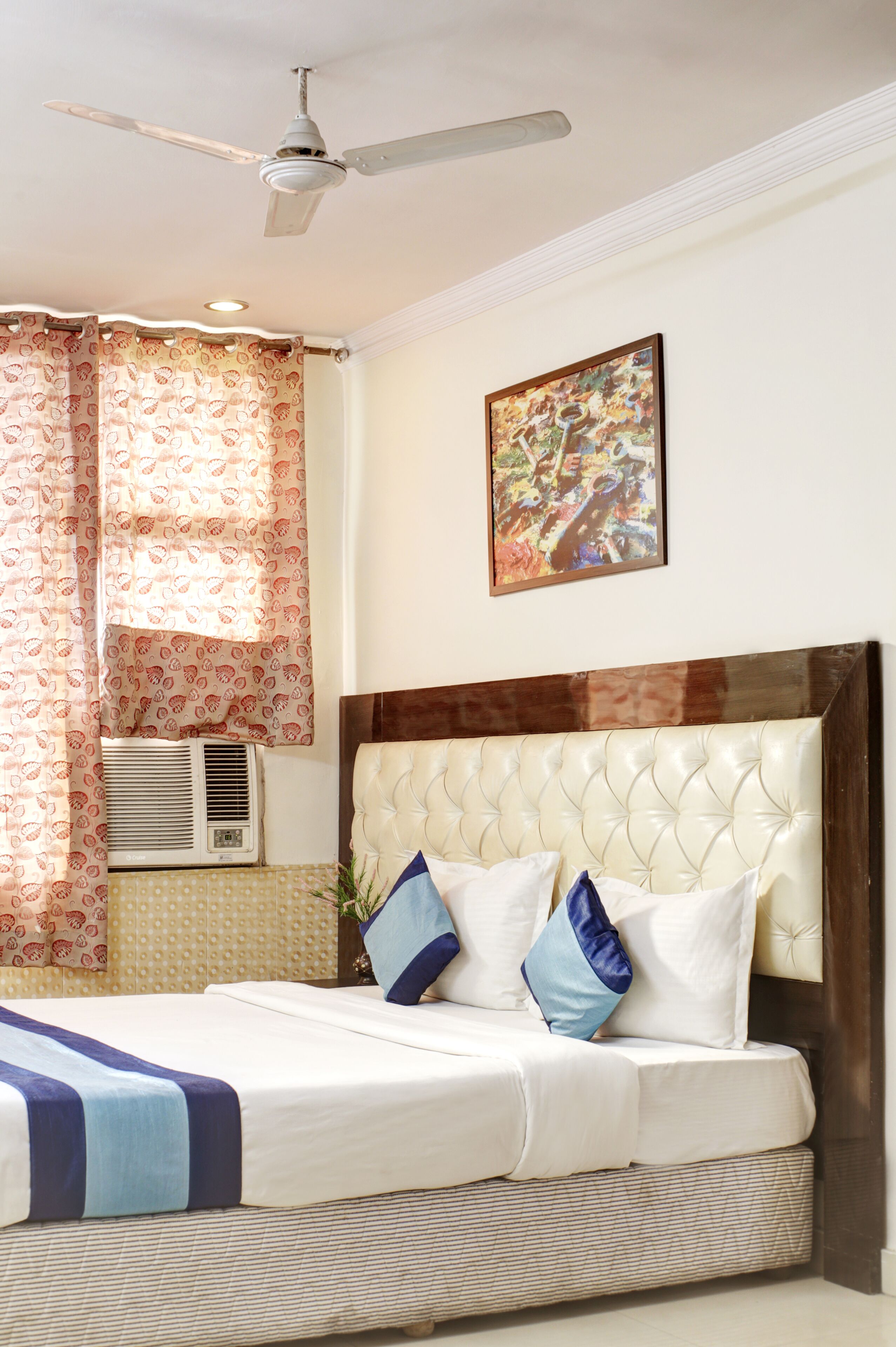 A hotel room with a bed, a ceiling fan, a window with curtains, an air conditioner, and a painting on the wall.
