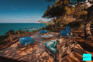 Eine Holzterrasse mit Sitzgelegenheiten im Freien, mit Blick auf eine Küstenlandschaft und freiem Blick auf das Meer und eine entfernte Insel.