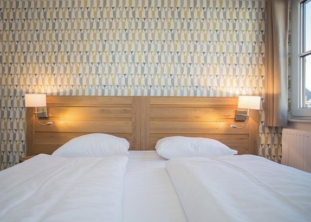 A double bed with white linens, wooden headboard, two bedside lamps, and a patterned wallpaper.