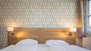 Cama doble con sábanas blancas, cabecero de madera, dos lámparas de noche y un papel tapiz estampado.