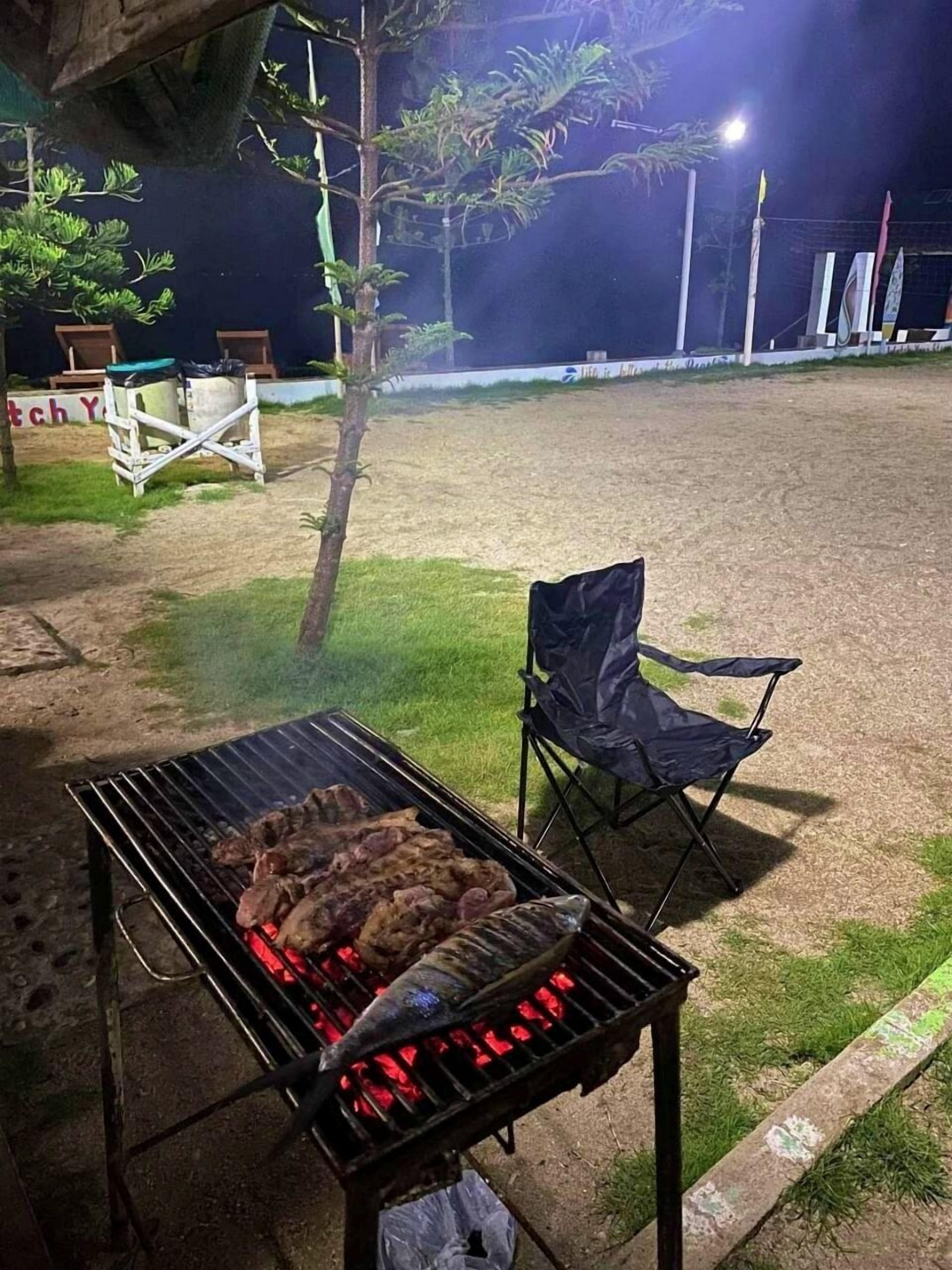 Una parrilla al aire libre con carne asándose, una silla plegable y un césped con una valla y árboles al fondo.