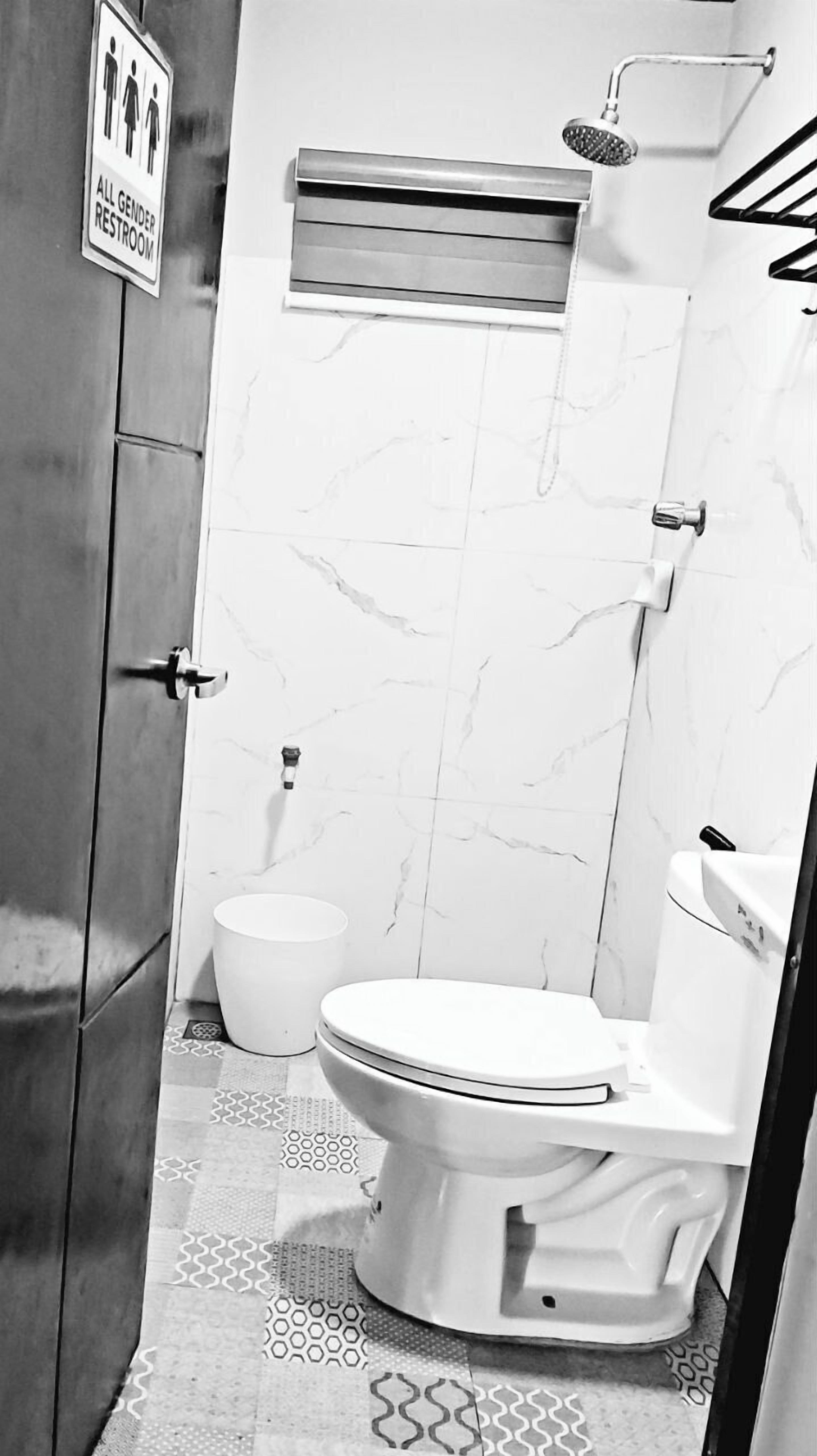 A bathroom with a toilet, a bidet, and a shower. There is a sign indicating it is an all gender restroom. The floor has a patterned tile design.