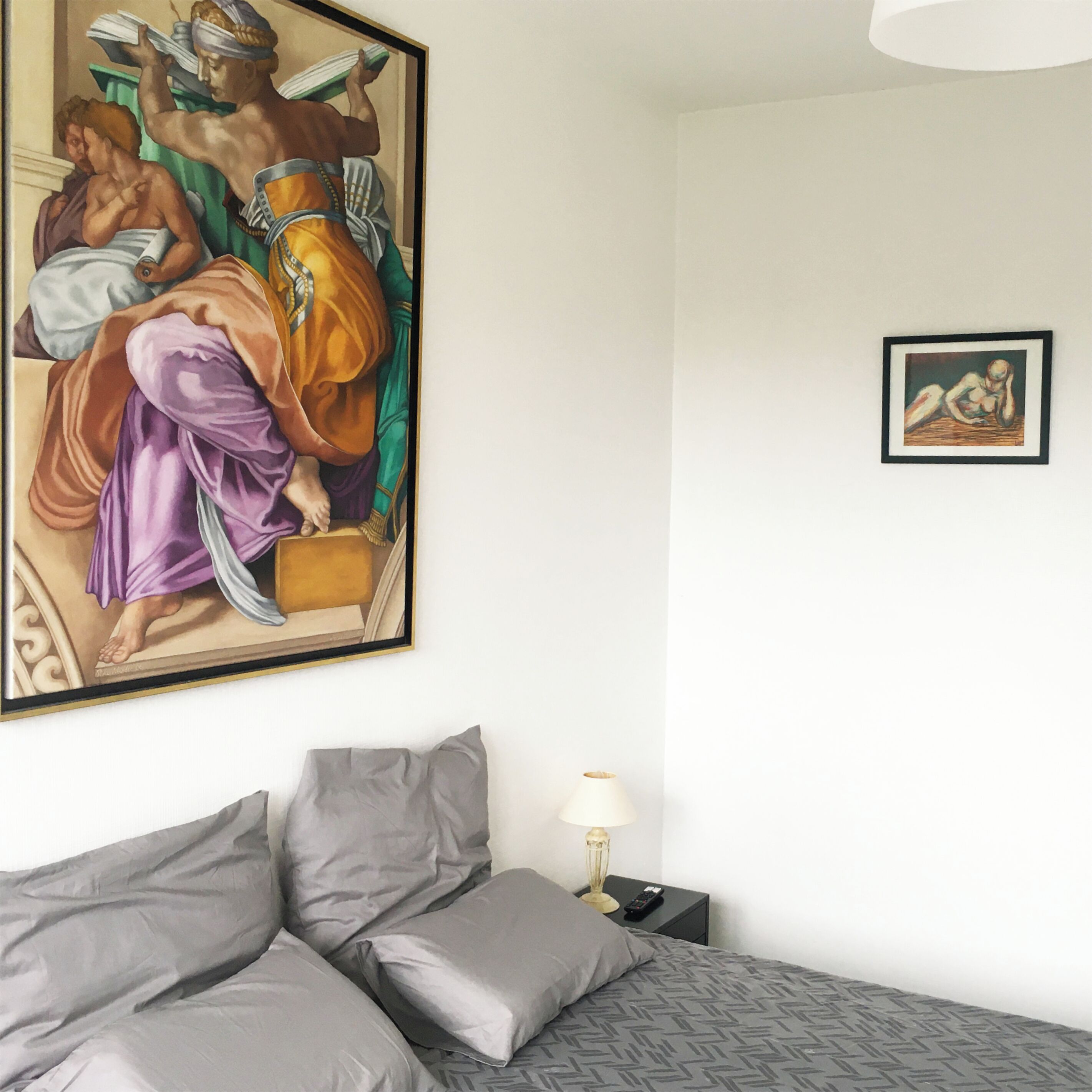Ein Schlafzimmer mit einem großen Gemälde im klassischen Stil, einem Bett mit grauer Bettwäsche, einem Nachttisch mit einer Lampe und einem kleineren gerahmten Kunstwerk an der Wand.