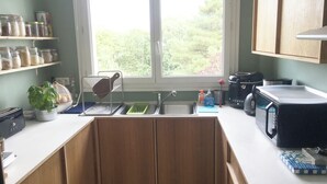 A kitchen with a window, wooden cabinets, a sink, and various appliances. - Juste une Chambre Chez L'habitant (Saint-Sébastien-sur-Loire)
