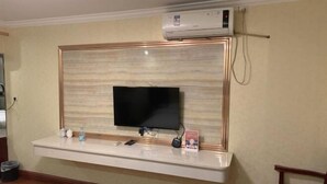 A flat-screen TV mounted on a wall with a wooden panel design, a small shelf below holding a water bottle, a remote, and a small decorative item. - Jixuan Hotel (Zhongshan)