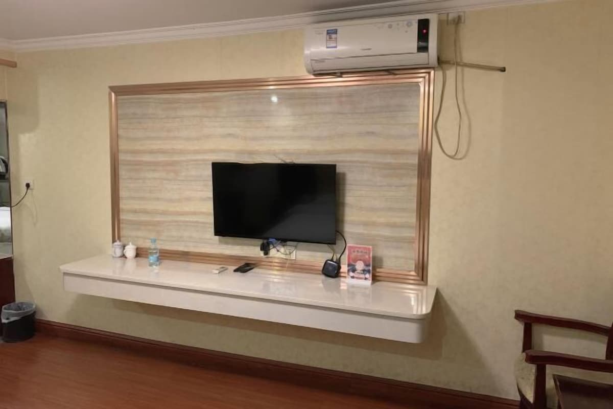 Un téléviseur à écran plat fixé au mur, avec un panneau décoratif en bois, et une petite étagère en dessous contenant une bouteille d’eau, une télécommande et un petit objet décoratif.