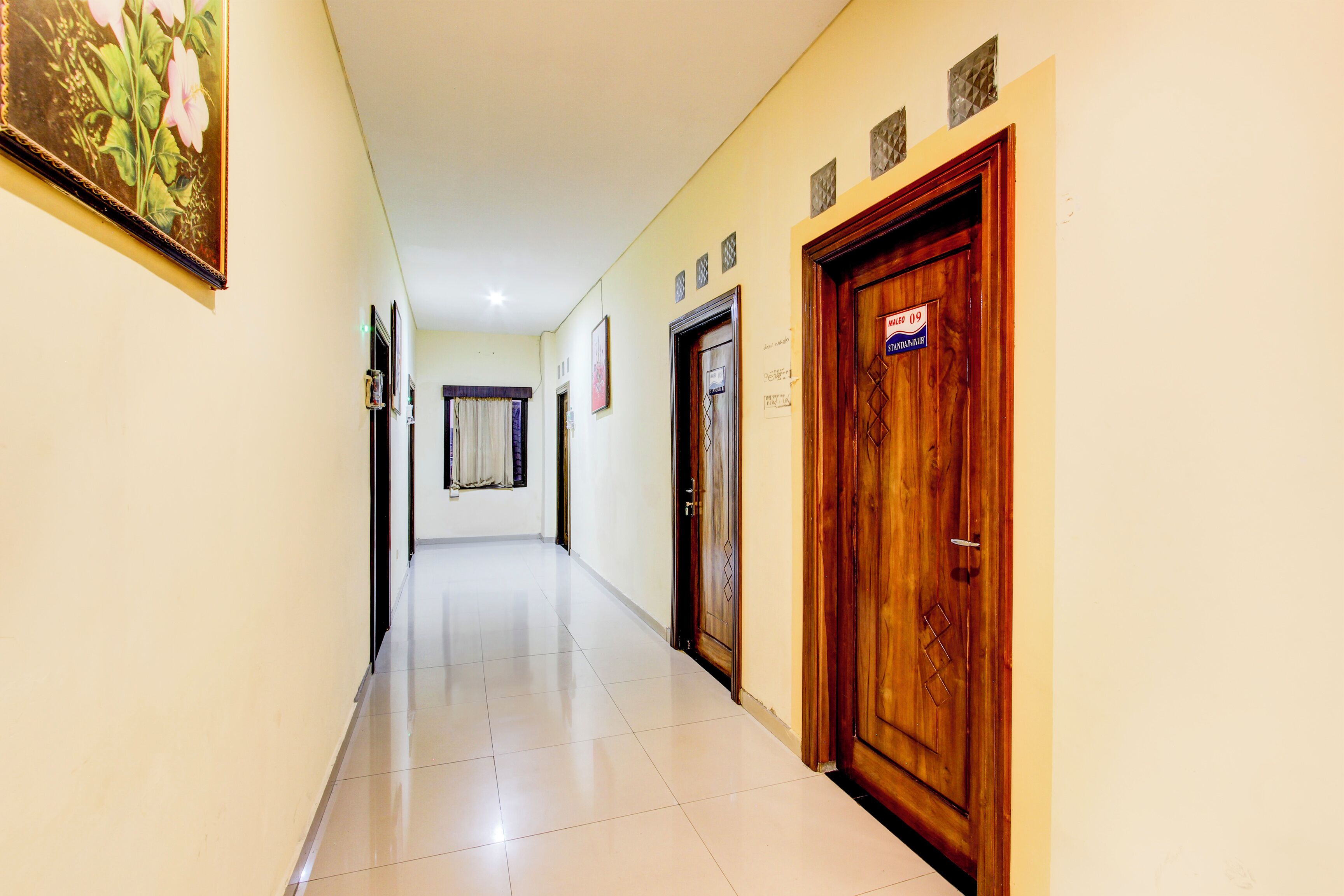 A hallway with a wooden door on the right, a framed picture on the left, and a window with curtains in the background.