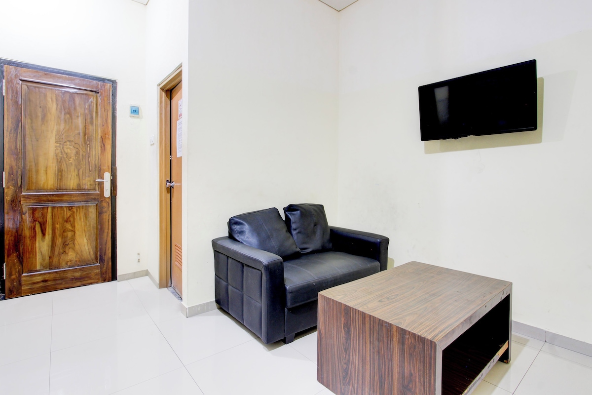 Una habitación con puerta de madera, un sofá de cuero negro con cojines, una mesa de centro de madera y un televisor de pantalla plana montado en la pared.