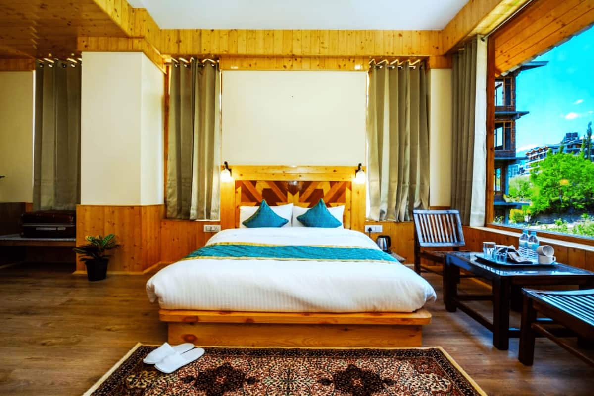 A wooden bedroom with a large bed, curtains, a window with a view of greenery, and a small table with a chair.