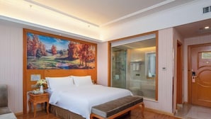 A hotel room with a large bed, a wooden headboard, a painting of a landscape, a bench, a nightstand with a vase of flowers, and a glass door leading to a bathroom. - Vienna Hotel (Dunhuang)