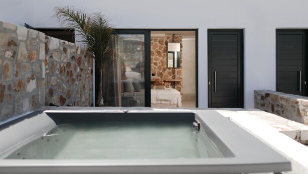 A modern outdoor pool area with stone walls, a glass door leading to a room with a bed and a mirror, and two black doors. - Delight Apartments Suites (Naxos)