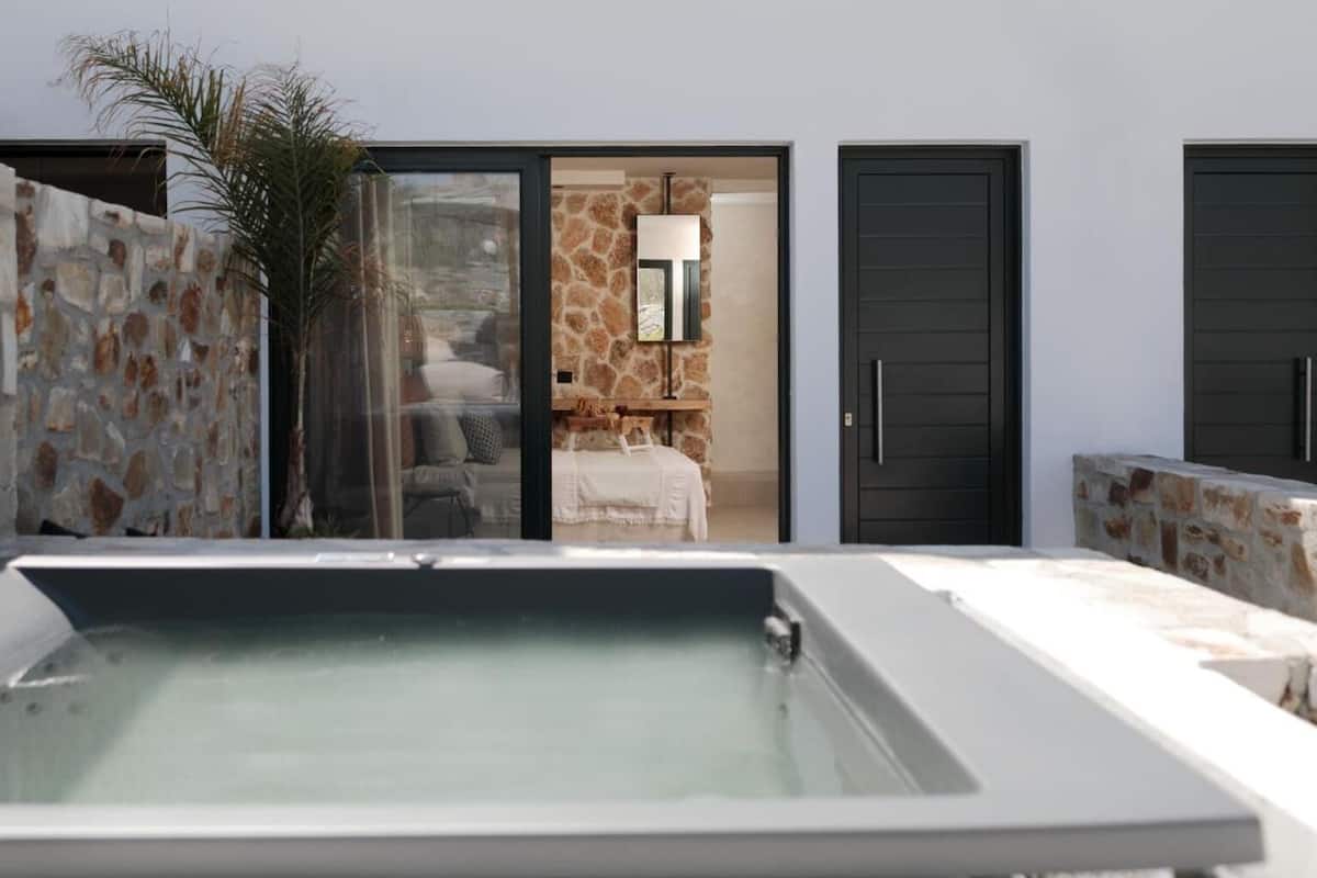 A modern outdoor pool area with stone walls, a glass door leading to a room with a bed and a mirror, and two black doors.