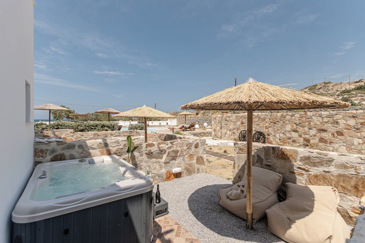 A stone-walled outdoor area with a hot tub, lounge chairs, and thatched umbrellas.