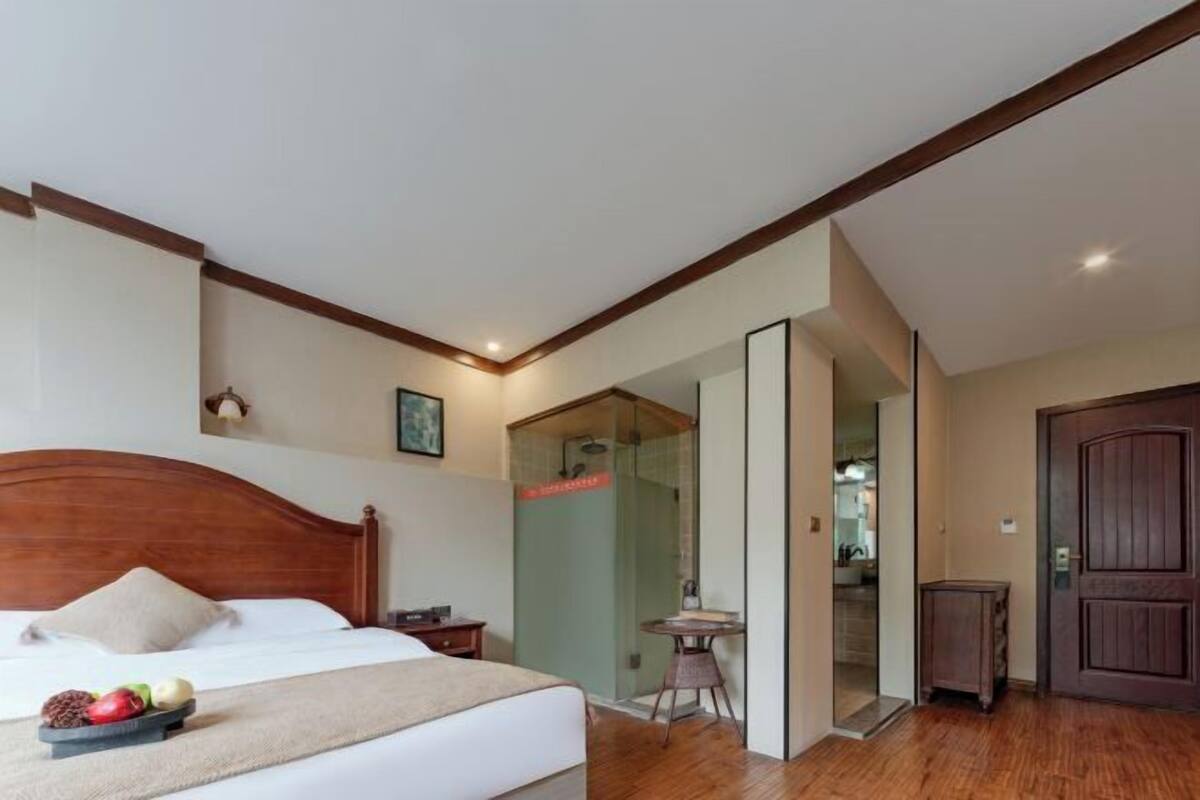 A hotel room with a wooden headboard, a bed with a white comforter, a small table with fruits, a glass-enclosed shower, and a wooden door.