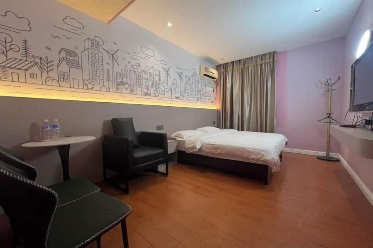 A hotel room with a bed, a chair, a small table, a coat rack, and a wall with a cityscape mural.