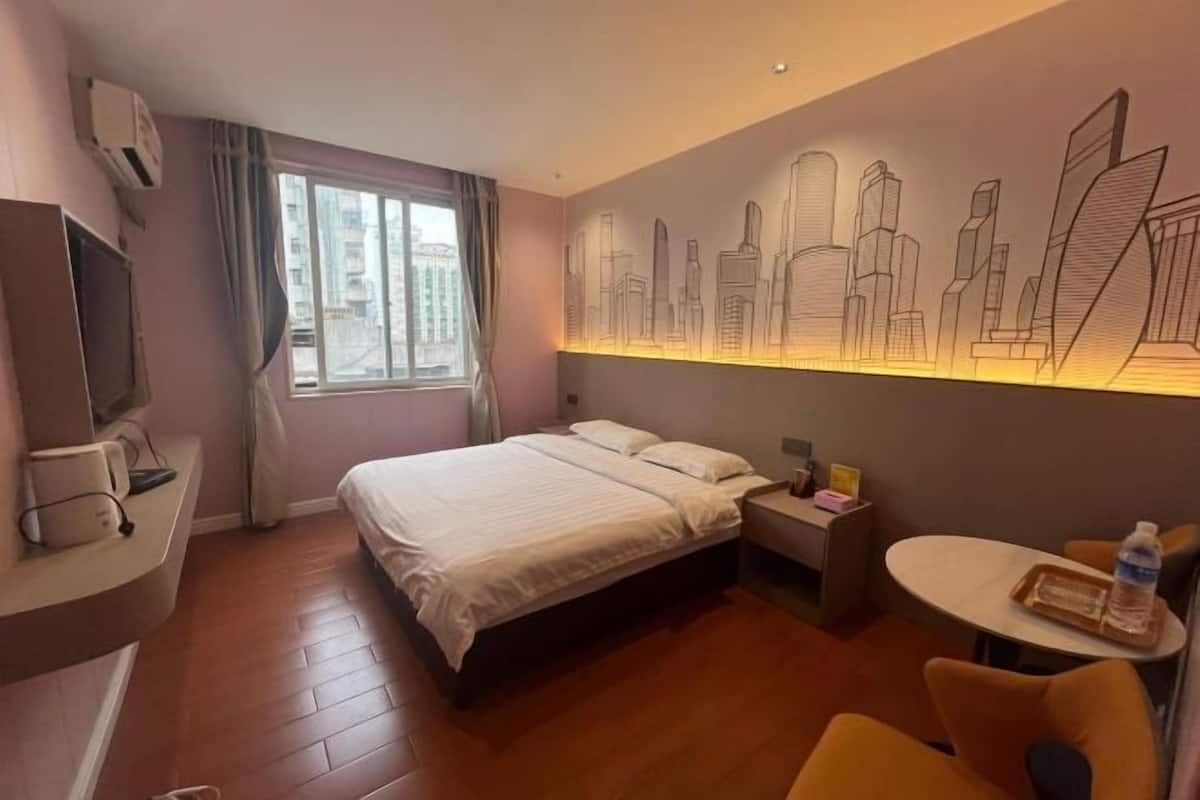 A hotel room with a bed, a desk, a chair, a table, and a wall with a cityscape mural.