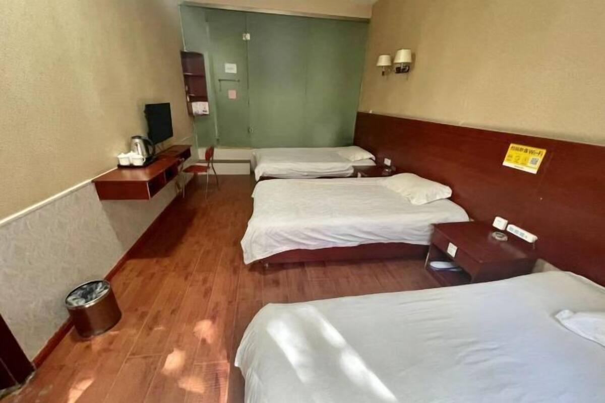 Quarto de hotel com três camas, piso de madeira, televisão, lixeira e luminária fixada na parede.