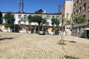 A paved courtyard with a car, trees, and multi-story buildings in the background.