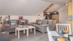 A modern living room with a grey sofa, white coffee table, and a kitchen area with wooden cabinets and a microwave.