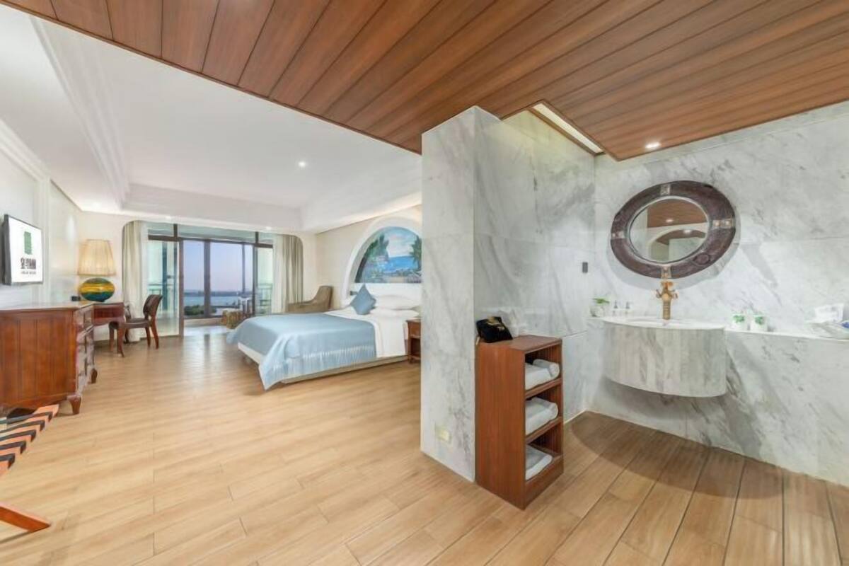 A hotel room with a wooden ceiling, a marble wall, a bed with a blue bedspread, a wooden nightstand, a marble sink, and a view of the outdoors through a large window.