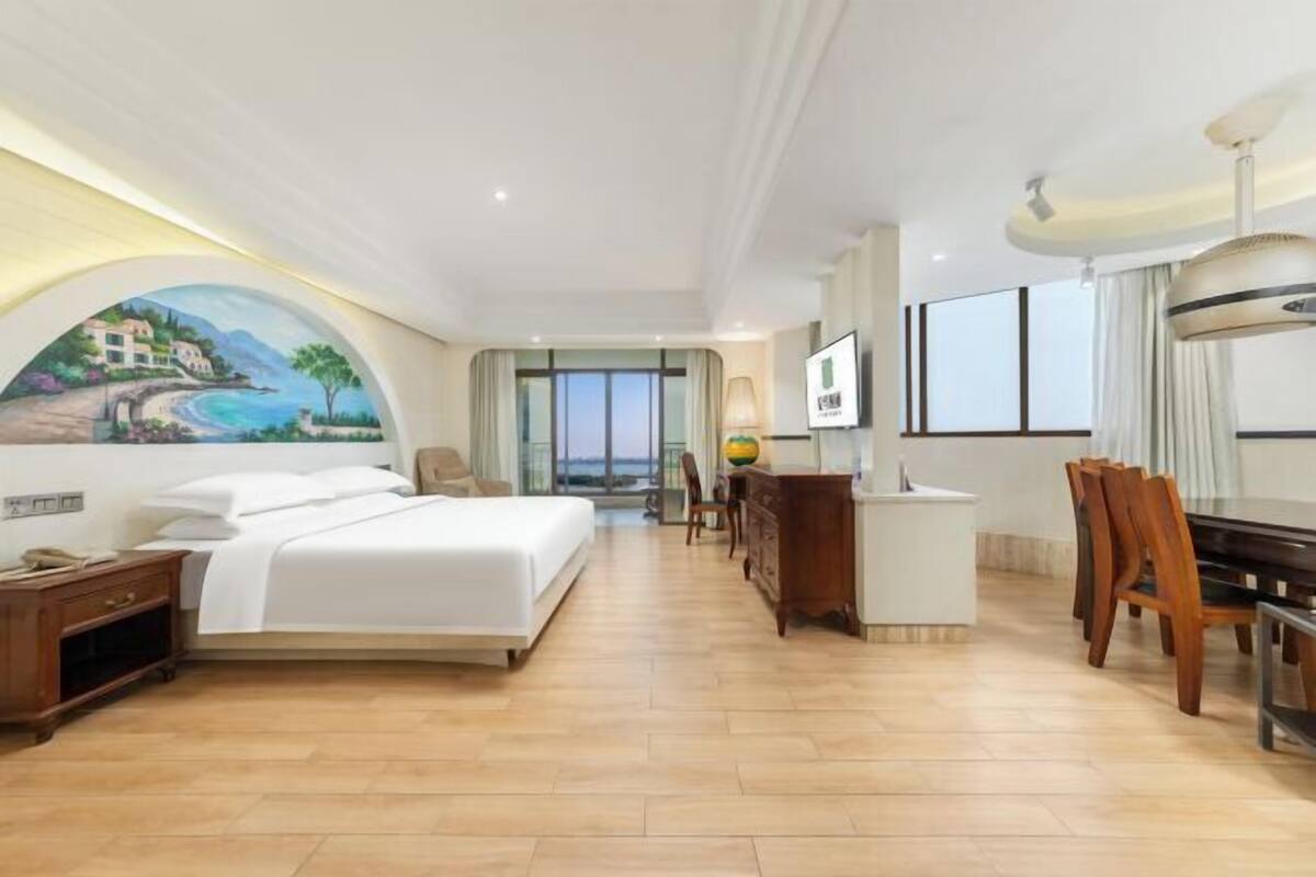 A hotel room with a bed, a desk, a chair, a TV, and a painting of a coastal scene.