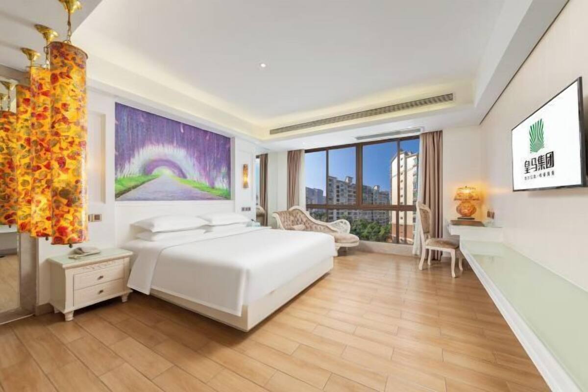 A hotel room with a bed, a nightstand, a chair, a large window, and a painting on the wall.