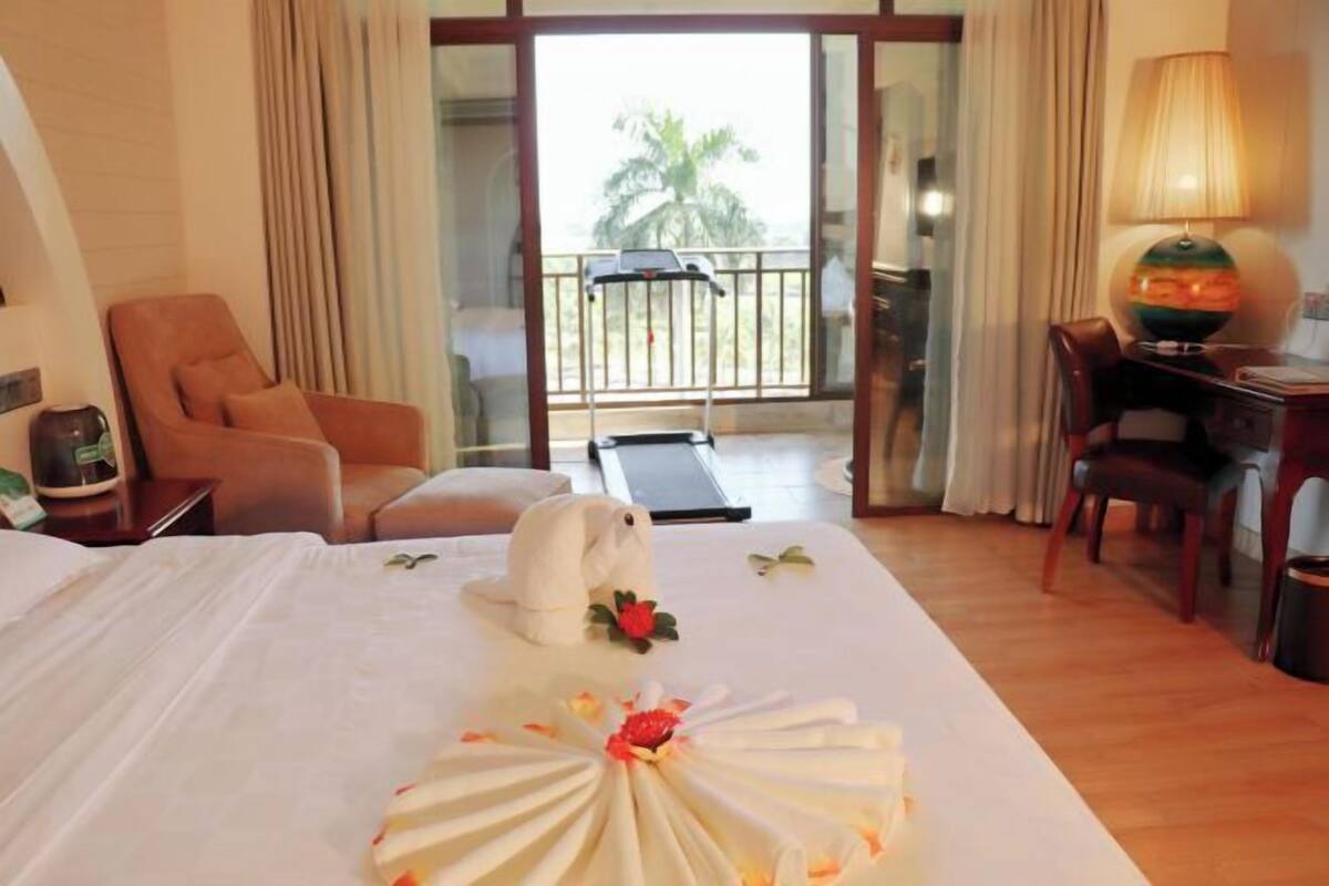 A hotel room with a bed, a chair, a desk, a lamp, and a view of a balcony with a palm tree.