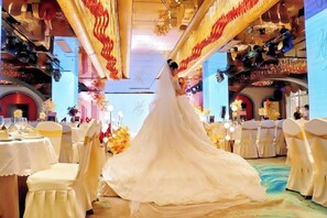 A wedding ceremony in a banquet hall with tables set for guests, floral decorations, and a bride in a white gown.