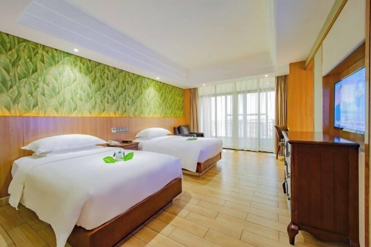 A hotel room with two beds, a wooden desk, a television, and a large window with curtains.