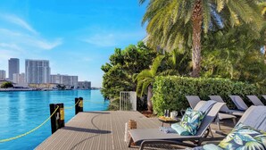 A poolside area with lounge chairs, a wooden deck, and a view of a city skyline across the water. - Amazing Apartment with pool,  Balcony, Near Beach (Hallandale Beach)