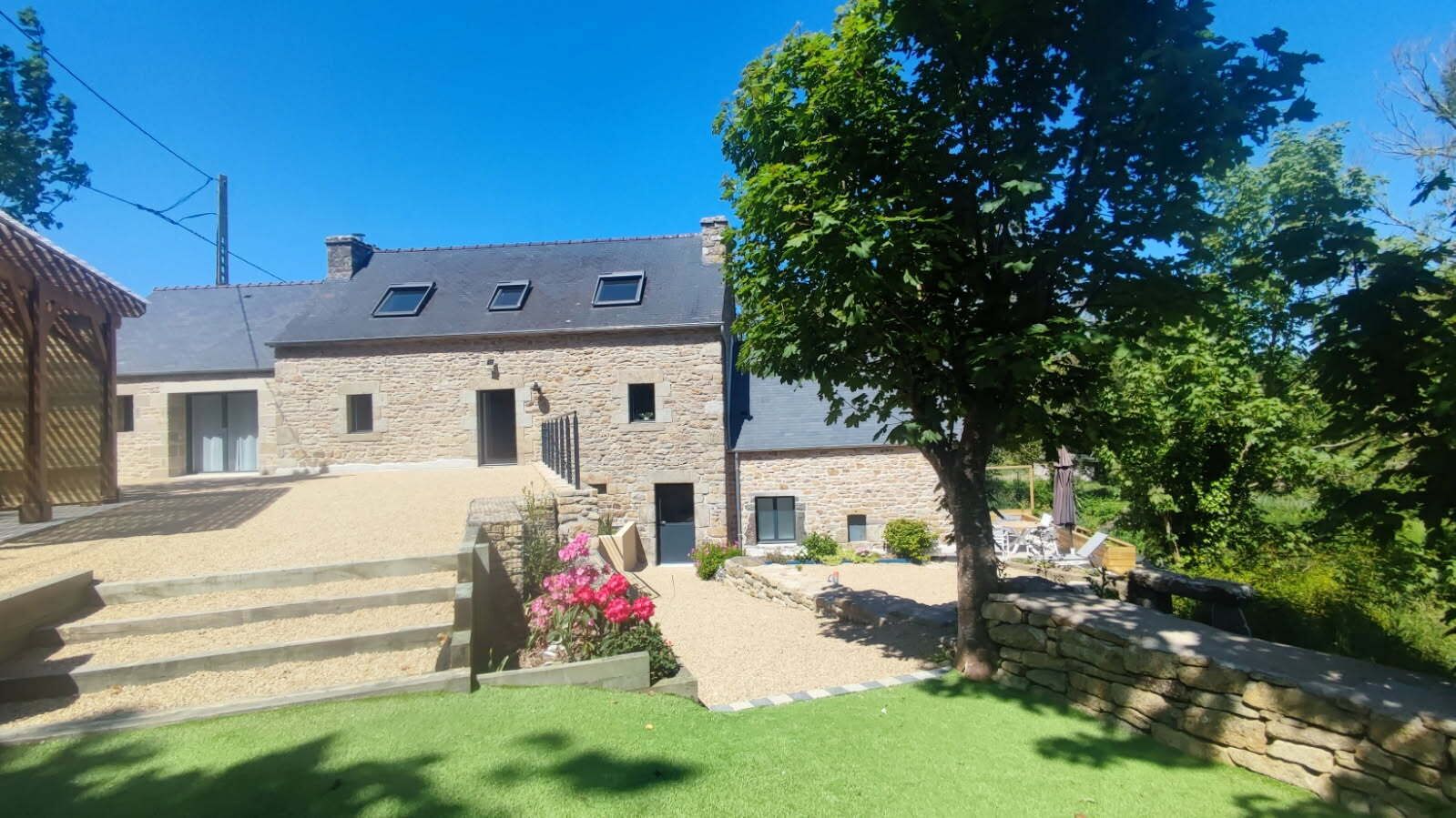 Un bâtiment en pierre avec un toit en pente, de nombreuses fenêtres et une allée pavée menant à un espace verdoyant agrémenté d’un arbre et d’un parterre de fleurs.