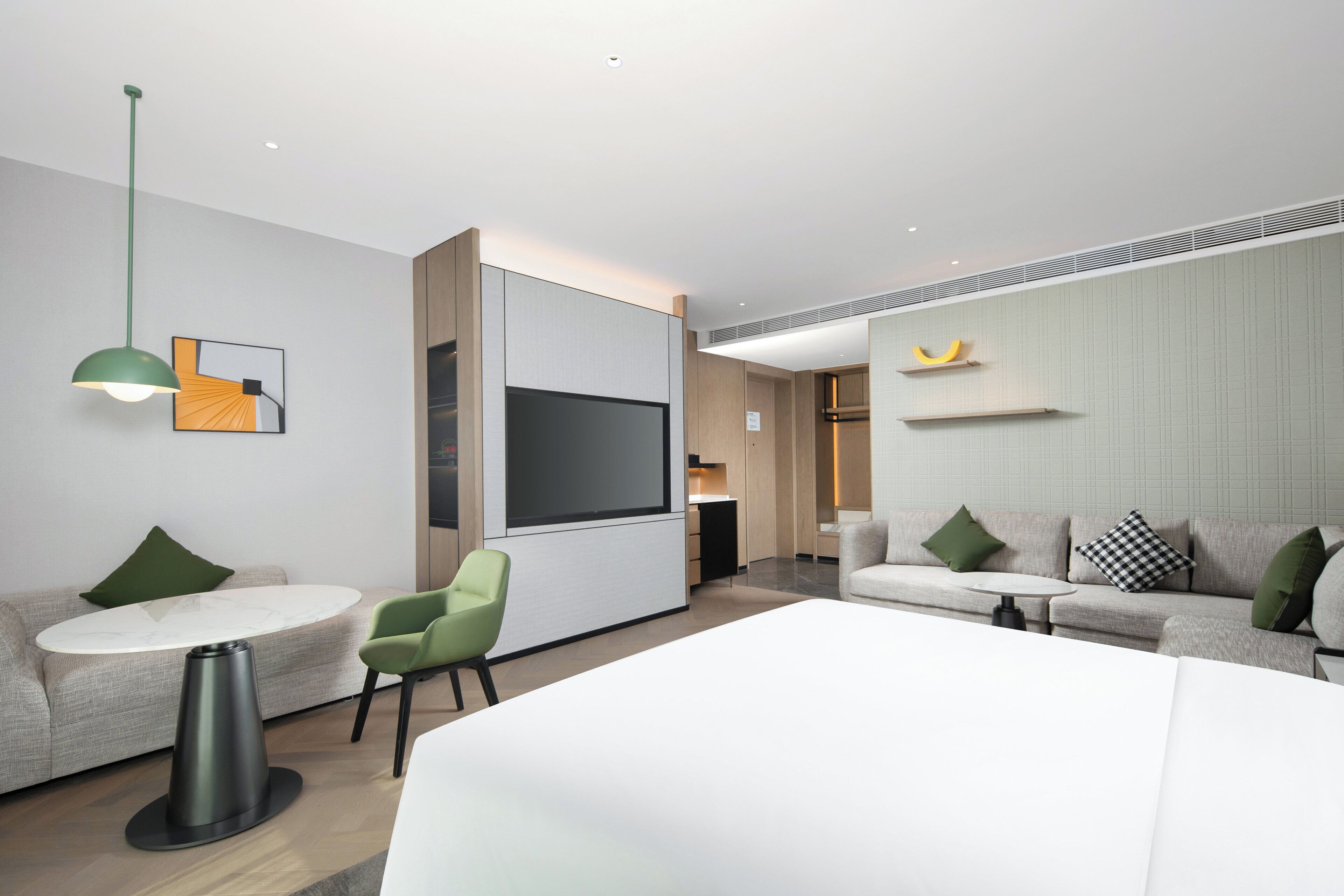 A modern hotel room with a large bed, two seating areas with tables, a flat-screen TV, and a green pendant light.