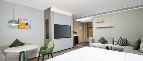 A modern hotel room with a large bed, two seating areas with tables, a flat-screen TV, and a green pendant light.