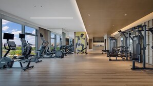 Egy modern edzőterem, melyben különféle edzőgépek, nagy ablakok és fa padló található.