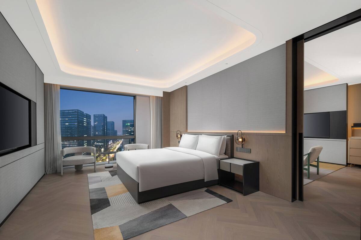 A modern hotel room with a large bed, a TV, a desk, and a city view.