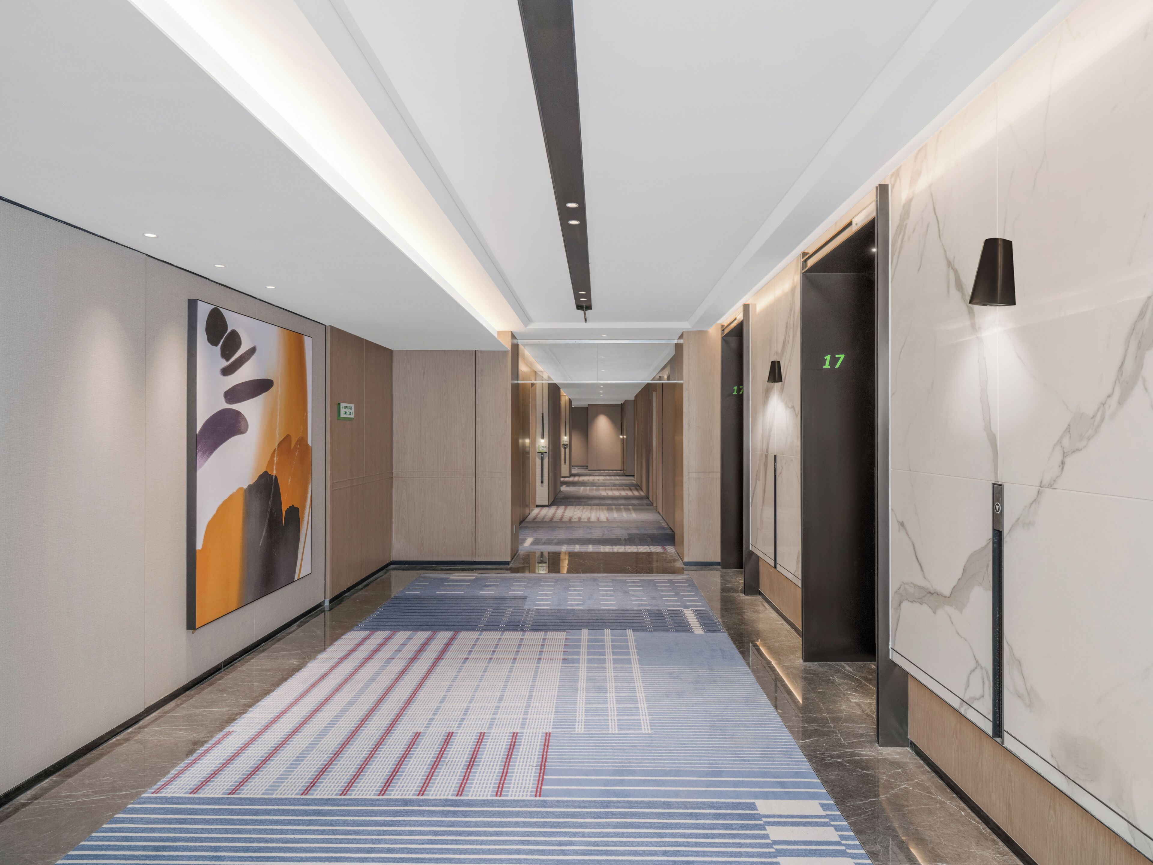 A modern hotel corridor with a long hallway, elevators, and a patterned carpet.