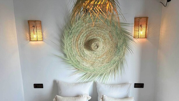 A bedroom with a bed, two white pillows, a woven wall hanging, and two wall-mounted lights.
