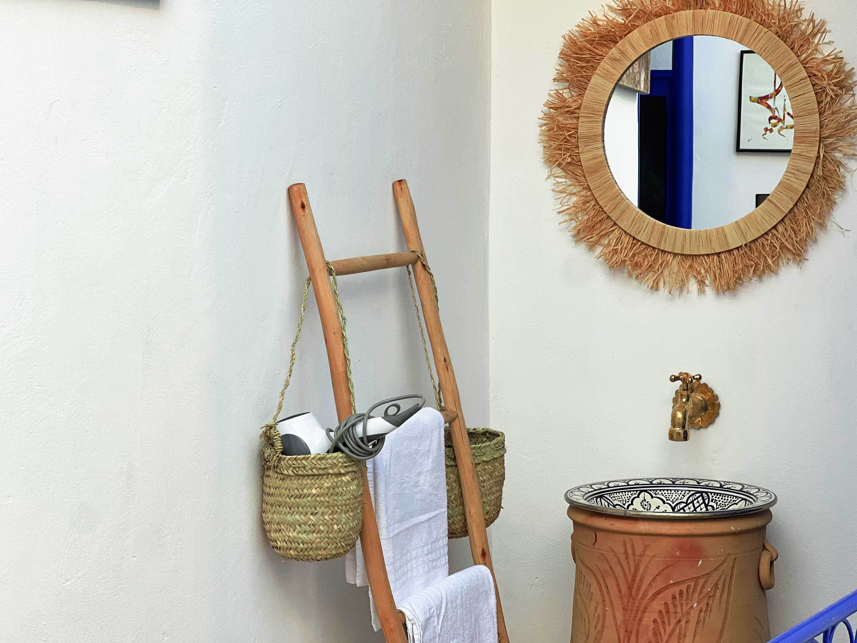Un baño con una escalera de madera apoyada en una pared, una cesta trenzada colgada en la pared, una toalla blanca colgando de la escalera, un espejo decorativo con marco de paja y una vasija de barro tradicional.