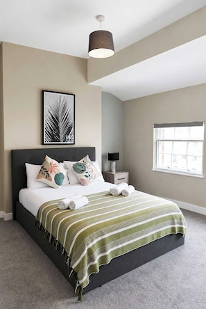 A modern bedroom with a bed, pillows, a bedside table, a lamp, a framed artwork, and a window.