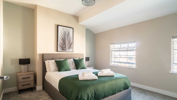 A bedroom with a large bed, a nightstand with a lamp, a framed artwork above the bed, and a window with blinds. - Charming 1 Bedroom Apartment in Central Liverpool (Liverpool)