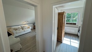 A room with a white sofa, a wooden door, and a window with a view of greenery. - Oberdeck - Friesische Gemütlichkeit (Wangerooge)