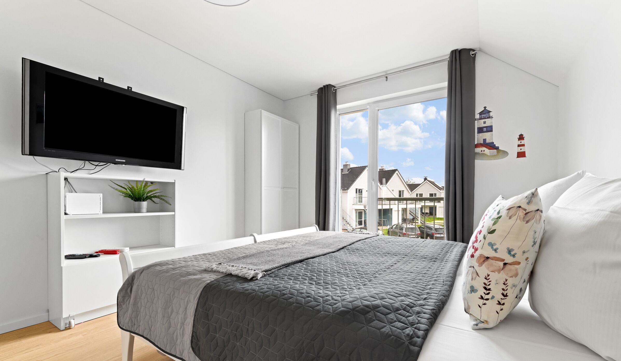 Une chambre moderne avec un grand lit, un téléviseur à écran plat, un bureau agrémenté d’une plante et une vue sur les maisons par la fenêtre.